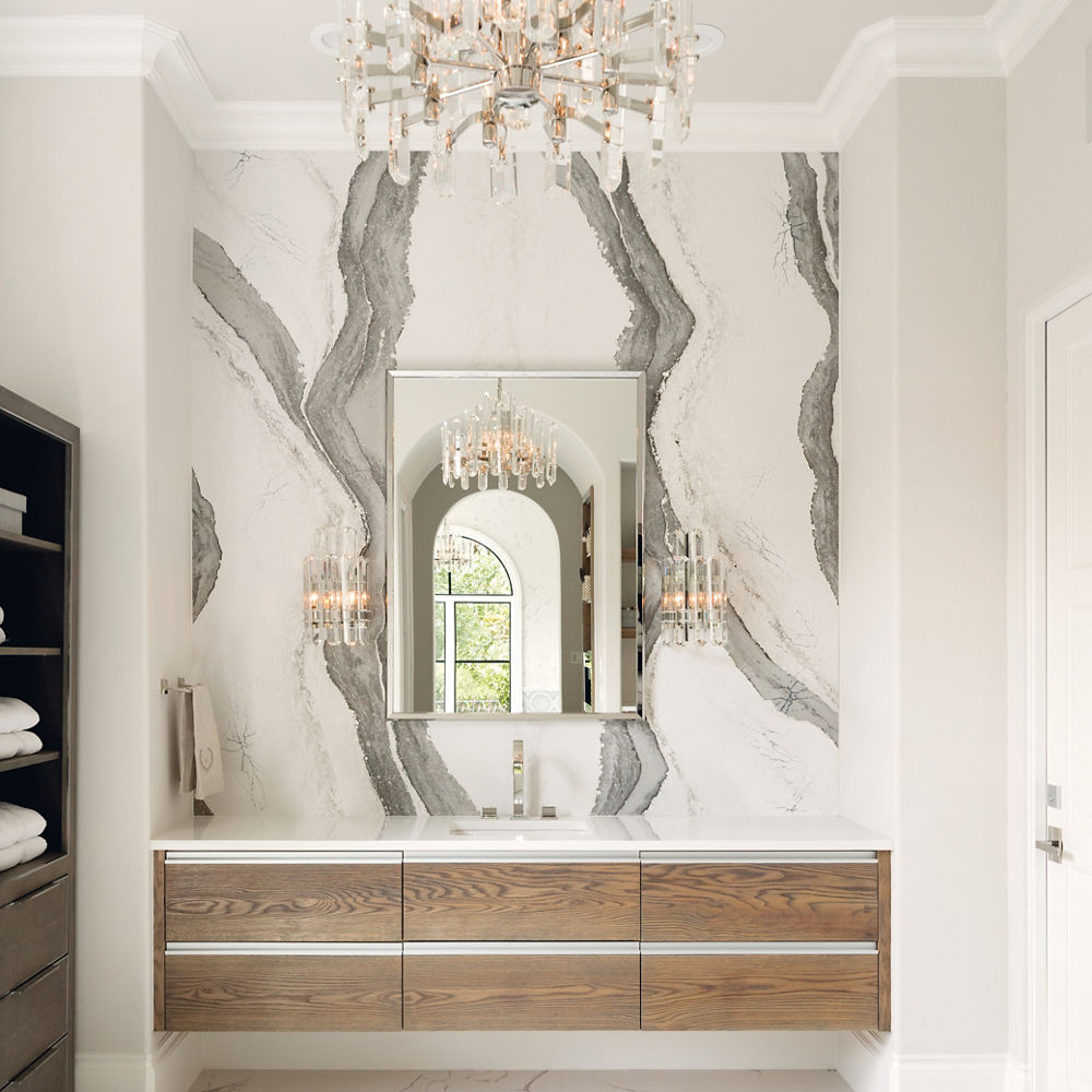 A gorgeous bathroom with grey and white veined quartz bookmatched backsplash, floating wooden vanity, silver mirror and chandelier, and lots of natural lighting. 