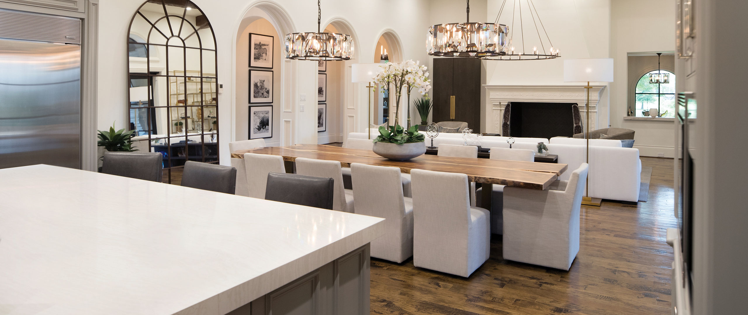 A gorgeous view from the kitchen of a dining table with two chandeliers, white cloth seating, and a living room in the background with a floor-to-ceiling fire place and overhead wooden beaming. 