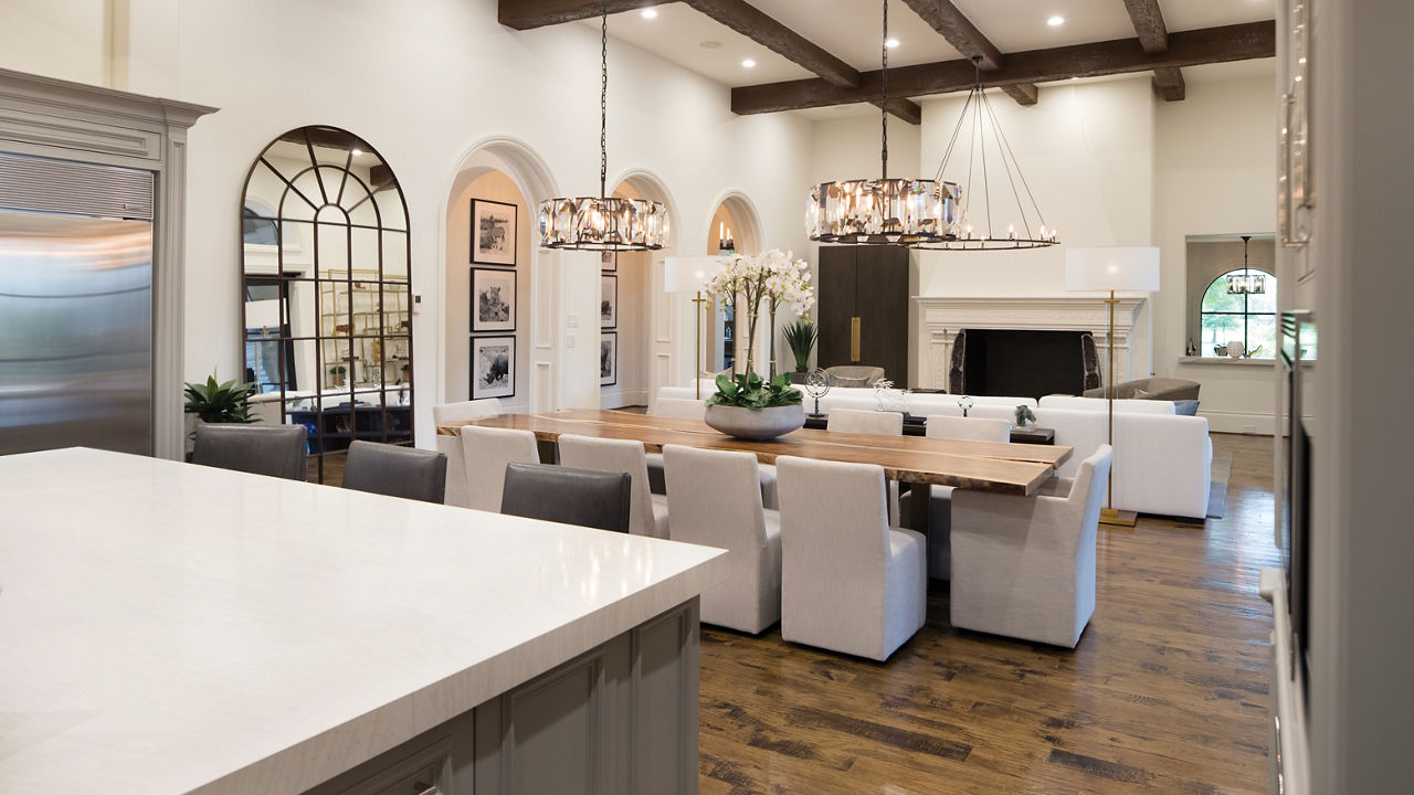 A gorgeous view from the kitchen of a dining table with two chandeliers, white cloth seating, and a living room in the background with a floor-to-ceiling fire place and overhead wooden beaming. 