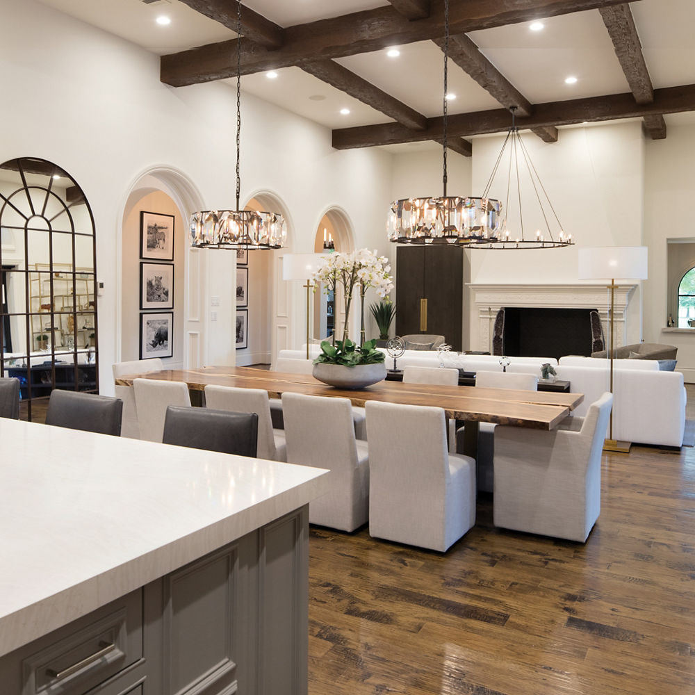 A gorgeous view from the kitchen of a dining table with two chandeliers, white cloth seating, and a living room in the background with a floor-to-ceiling fire place and overhead wooden beaming. 