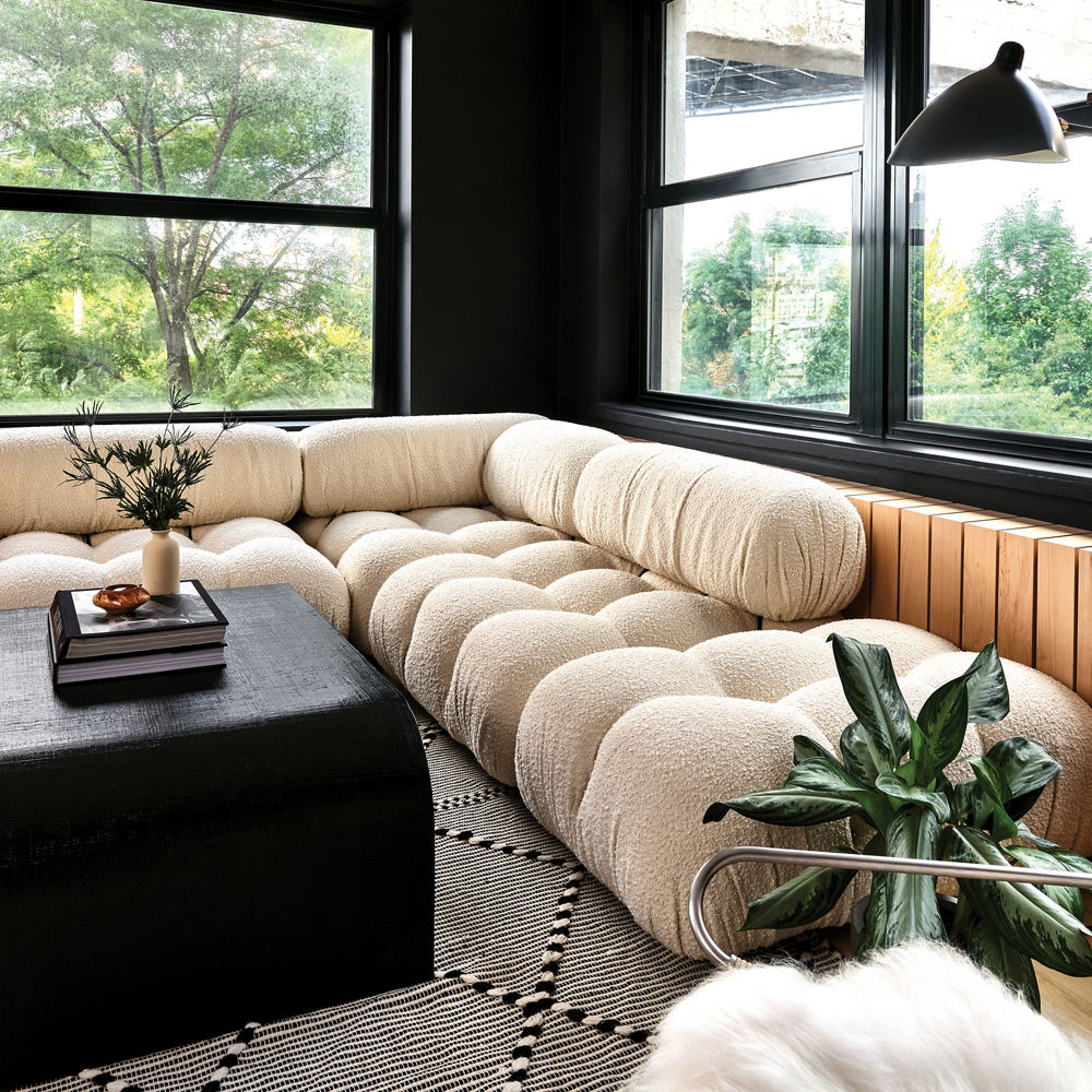 a cream, puffy sofa with a black ottoman, walls painted black with large black windows, and a hidden radiator covered in stylish wood.