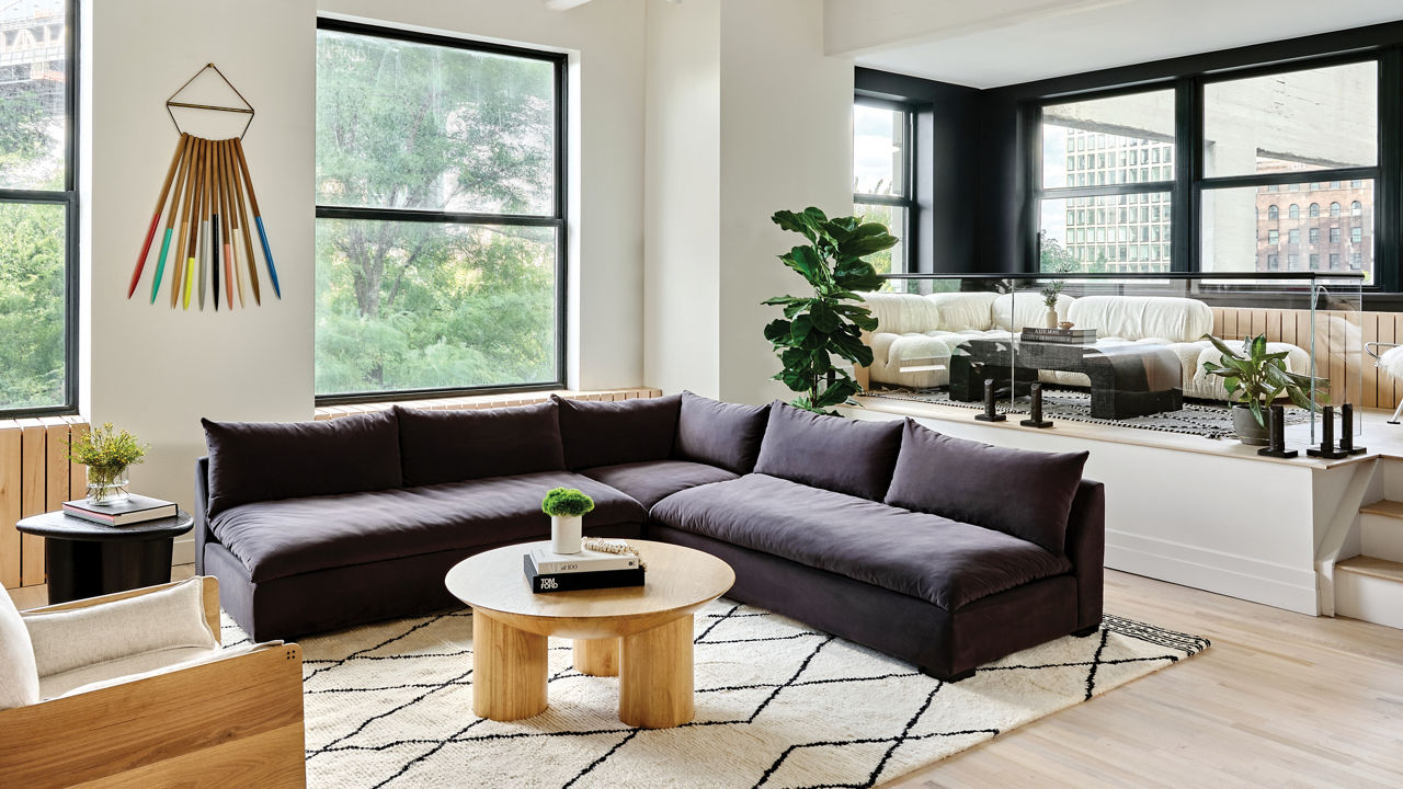 a gorgeous modern living room with a black sectional sofa, a wooden ottoman, light stained wooden floors and large black windows