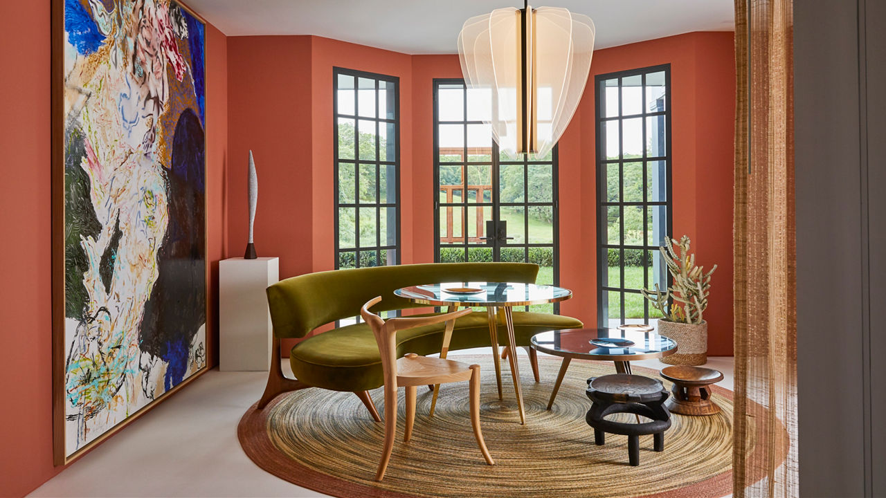 a bold dining room with burnt orange walls, a green bench, two tables, and a full-length wall painting.