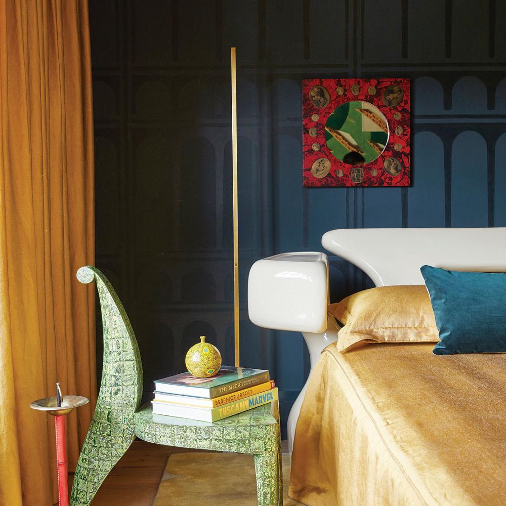 a bold bedroom with a dark blue accent wall, bed with white porcelain head frame, gold bedding, and a bold green chair with books on top.