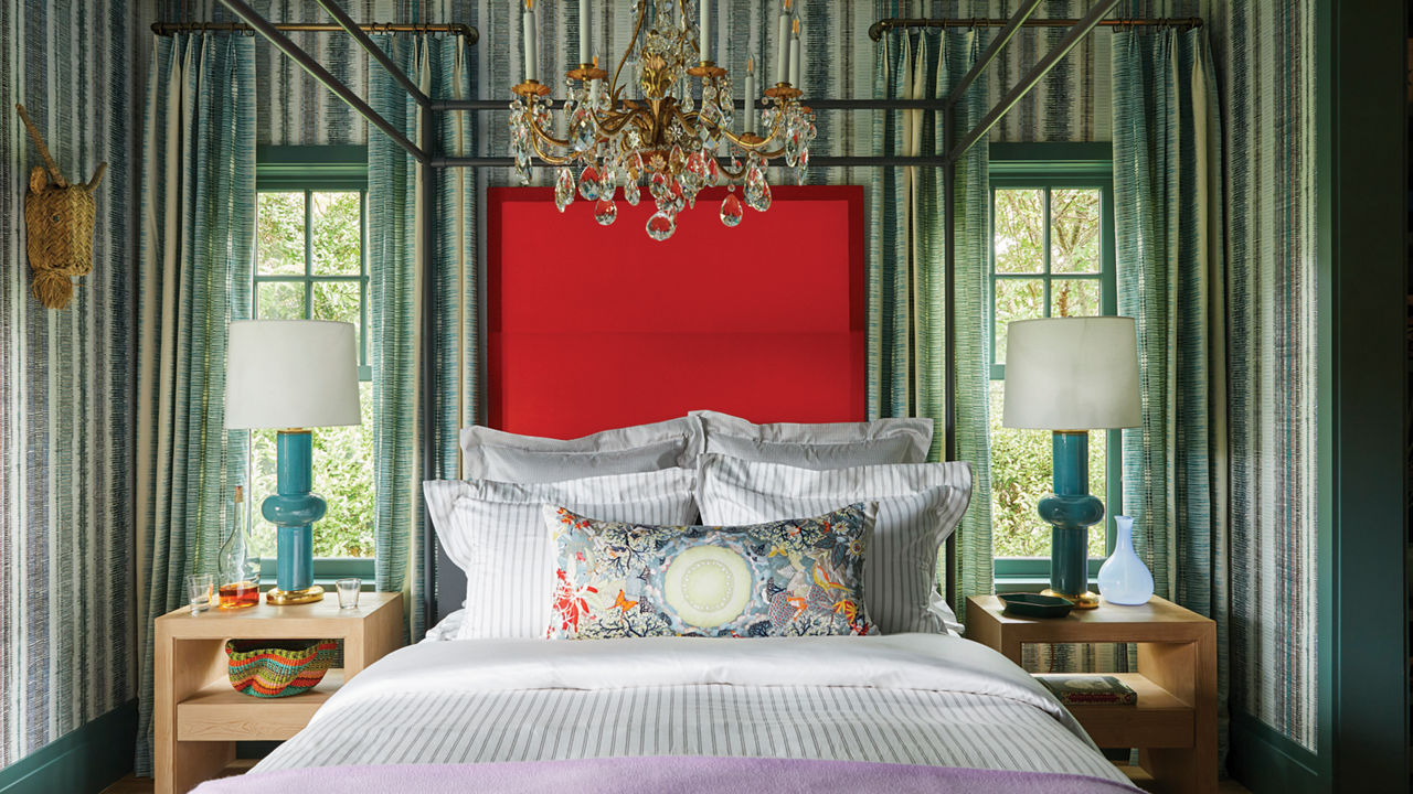 a bedroom with bold striped wallpaper, a crystal chandelier, nightstands with a teal and white lamp on top, and a red painting behind the bed. 