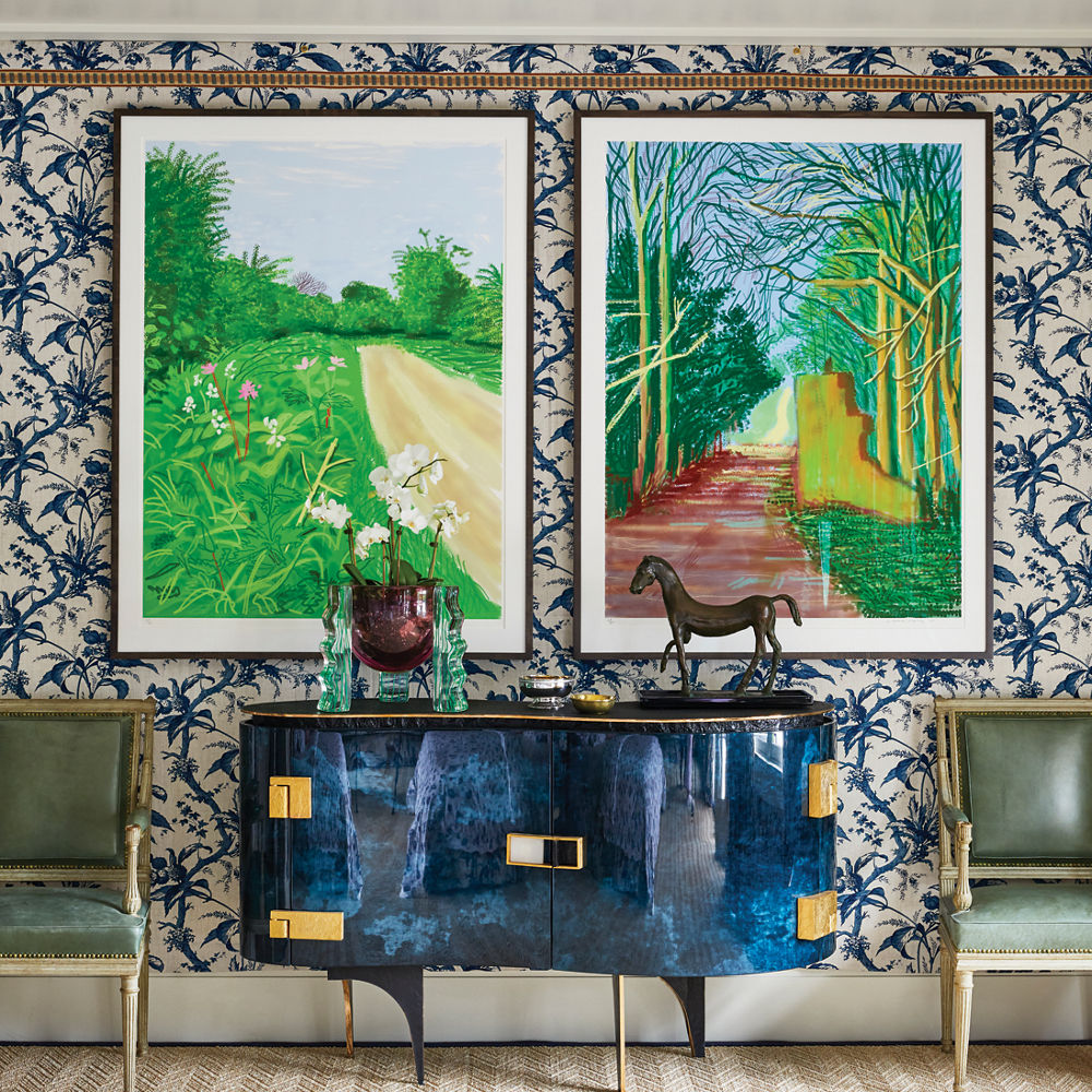 an entryway with boldly patterned wallpaper, a lapis buffet with two green chairs beside it, and two colorful paintings hanging side by side. 