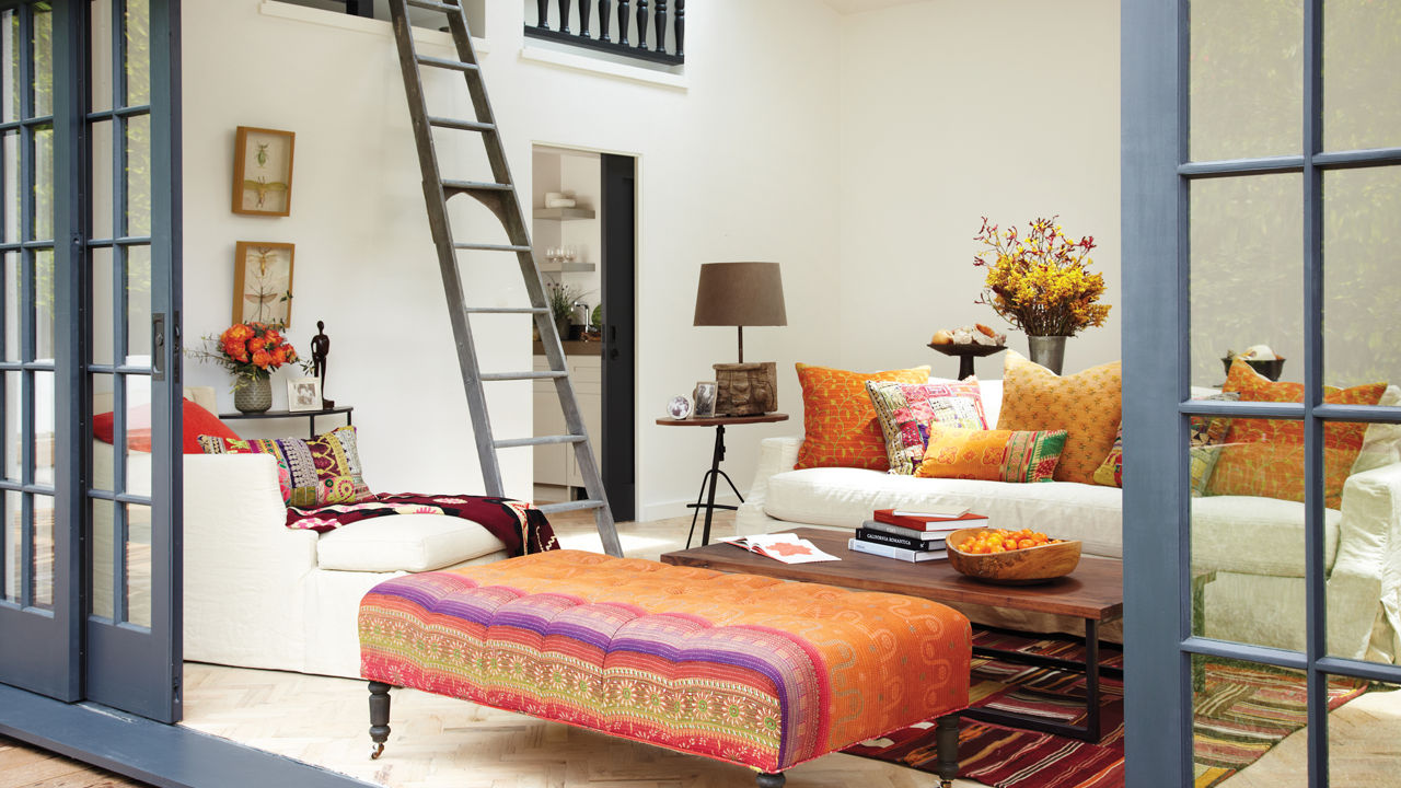 A living room with gorgeous blue french doors, and white furniture accented with orange and pink pillows and ottoman.