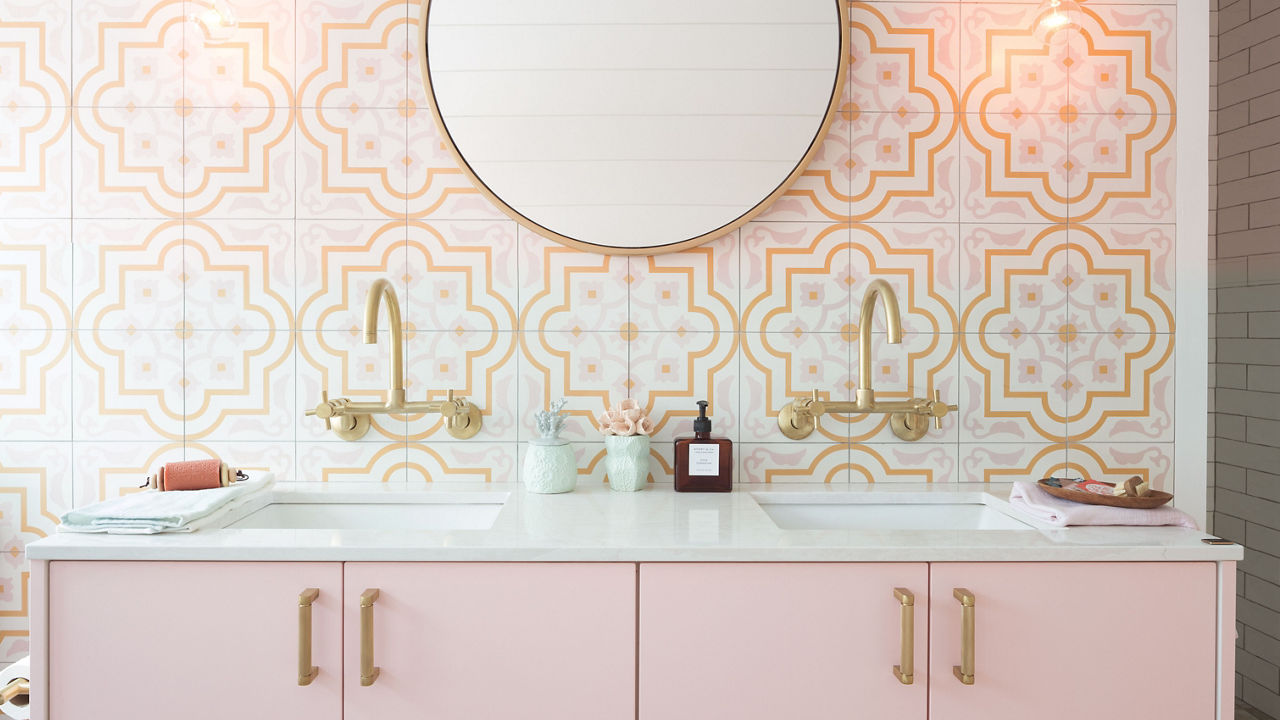 a cute bathroom with light pink cabinets, fun tiled backsplash, white quartz countertops, a round mirror, and gold hardware.