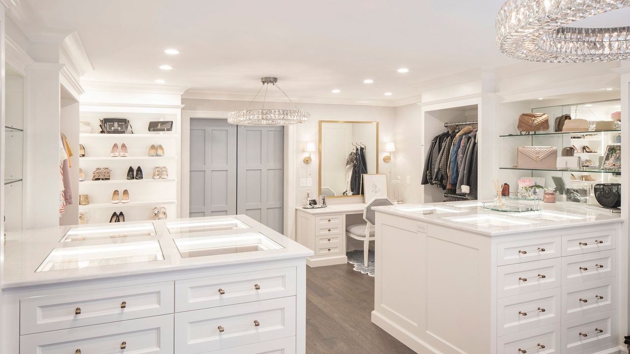 A modern and luxurious closet with two islands topped with white quartz countertop, open shelving for shoes and purchases, a vanity with a mirror and chair, and backlit lighting.