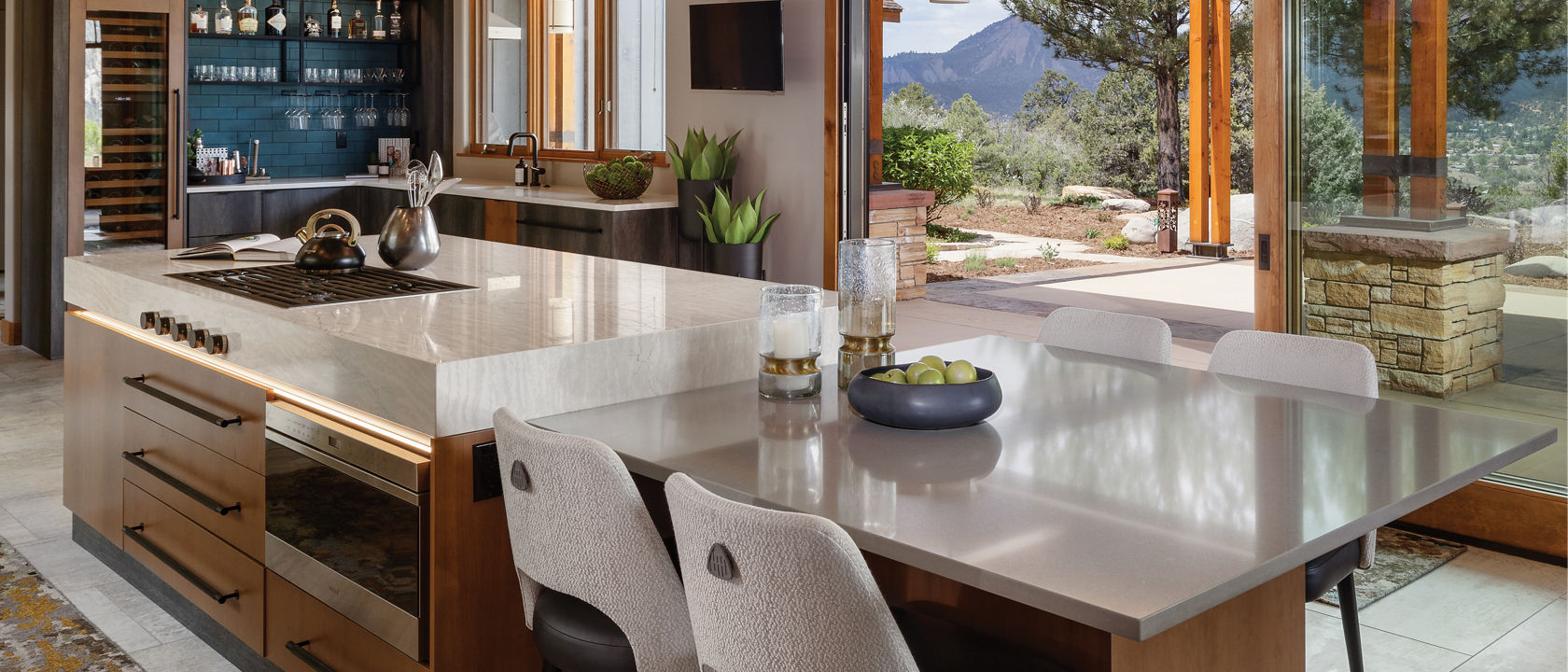 A warm and moody kitchen island with a modern built in stove and hood in the island, pull out drawers from the oak cabinets, side table next to the island with two white chairs, and a gorgeous view of the mountains and patio area. 