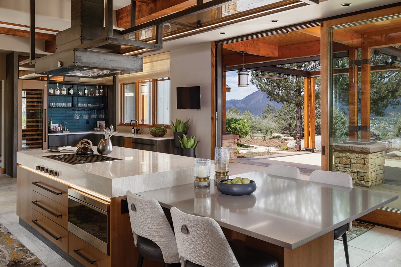 A warm and moody kitchen island with a modern built in stove and hood in the island, pull out drawers from the oak cabinets, side table next to the island with two white chairs, and a gorgeous view of the mountains and patio area. 