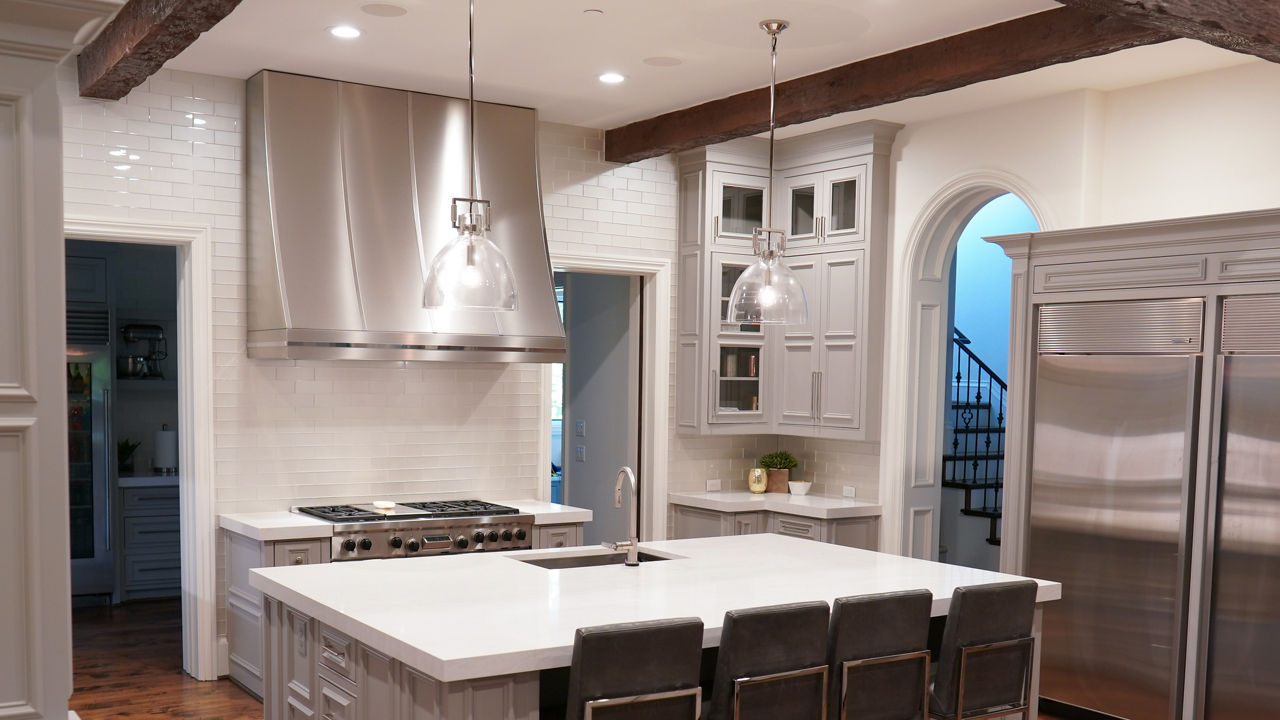 a traditional kitchen with light gray cabinets topped with white quartz countertops, an industrial style fridge, subway tile, and exposed ceiling beams. 