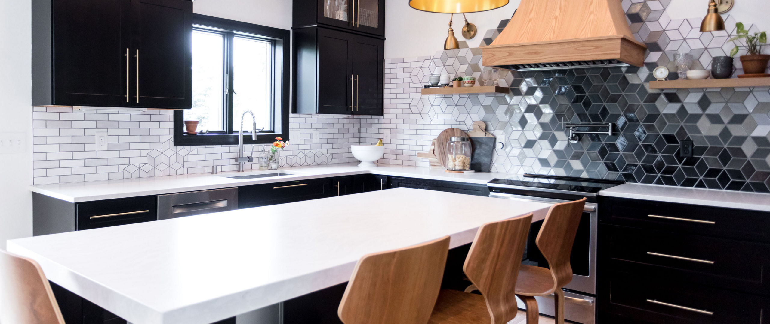 a black and white kitchen with black cabinets topped with white quartz countertops, a center island with 4 wooden barstools, a wooden hood over the range, and a black and white ombre hexagonal backsplash