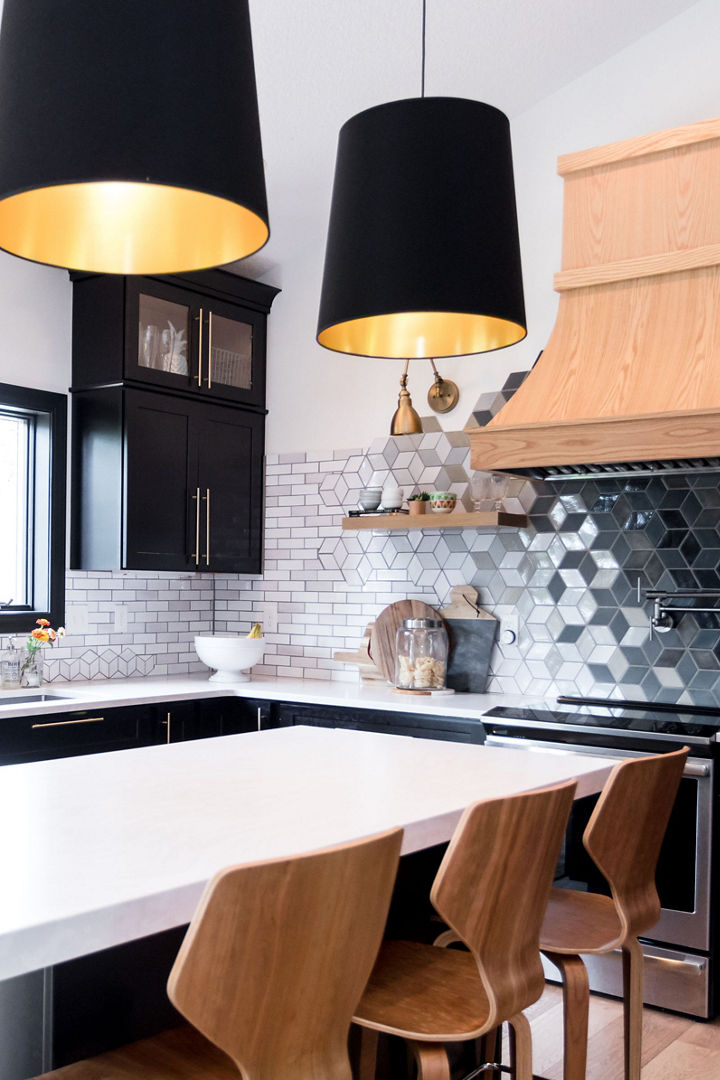 a black and white kitchen with black cabinets topped with white quartz countertops, a center island with 4 wooden barstools, a wooden hood over the range, and a black and white ombre hexagonal backsplash