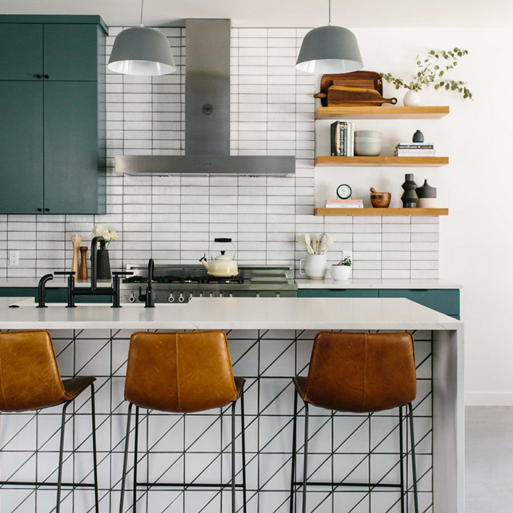 A blue-green kitchen with center double-waterfall edge island made from quartz countertops, blue-green cabinets, tile backsplash, and modern hood over range.