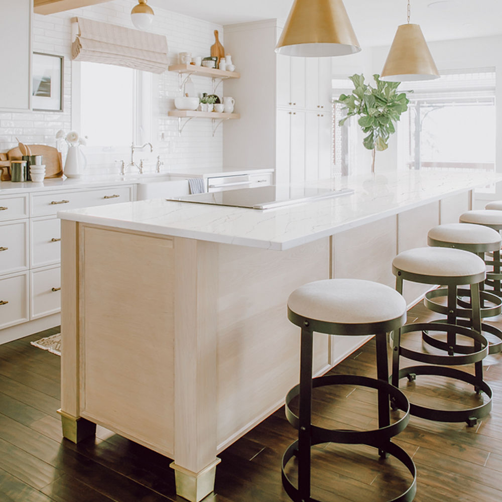 A kitchen with stained white oak cabinets topped with Cambria Quartz Ella countertops with brass accents in the ceiling beam and hanging ceiling lights.