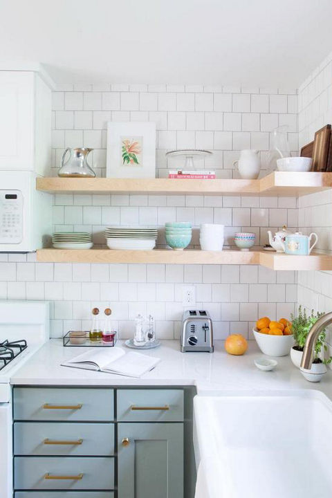 a galley kitchen with light green cabinets, farmhouse sink, open wooden shelving, subway tile, and one window with wood trim giving lots of natural light