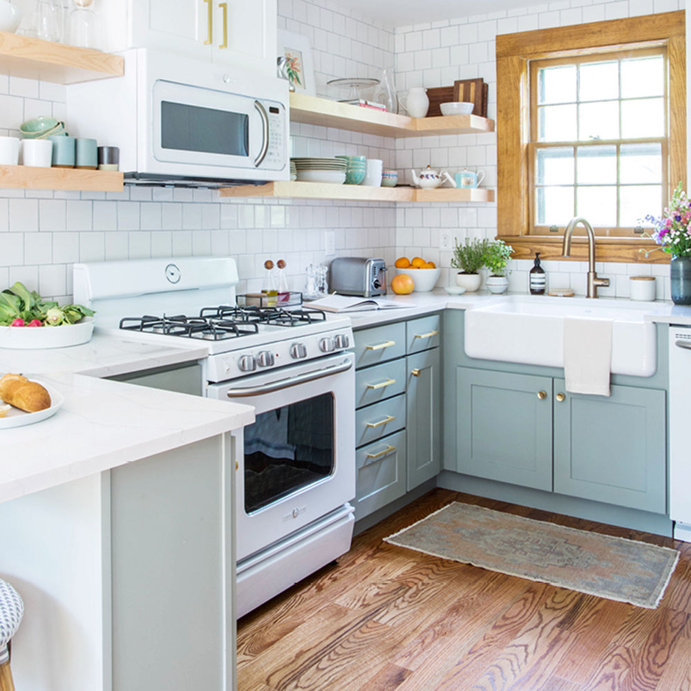 a galley kitchen with light green cabinets, farmhouse sink, open wooden shelving, subway tile, and one window with wood trim giving lots of natural light