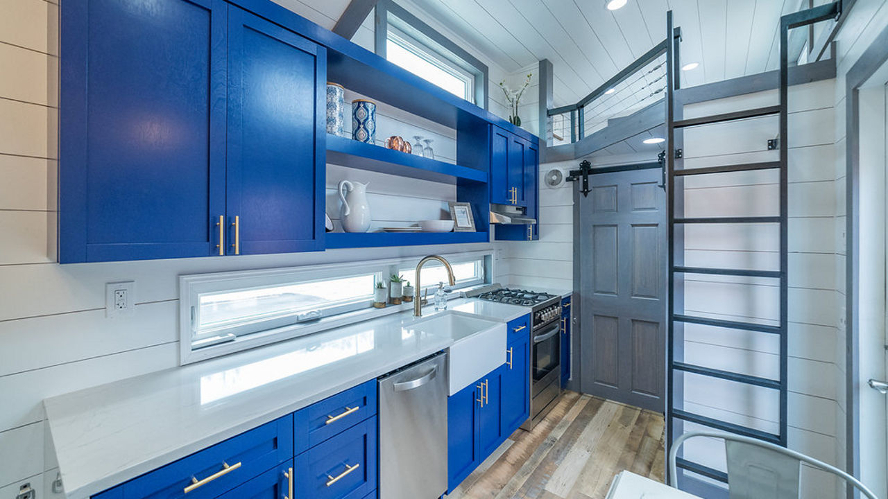 a tiny home with bright blue cabinets, white quartz countertops, open shelving, a blue door, trim, and ladder, and white cladding walls,