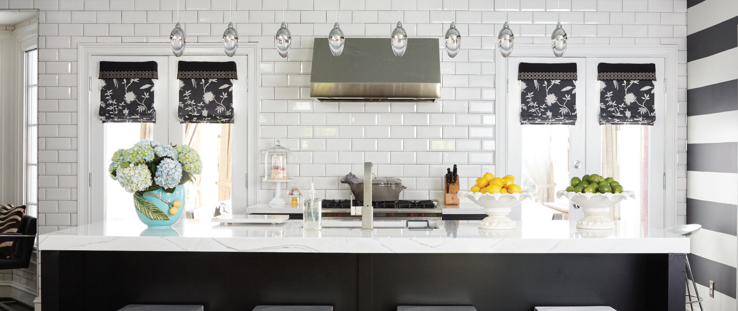 a black and white kitchen with a black center island topped with white quartz countertops and 4 white barstools, white subway tile backsplash, a black and gold range and hood and a black and white striped wall to the left. 