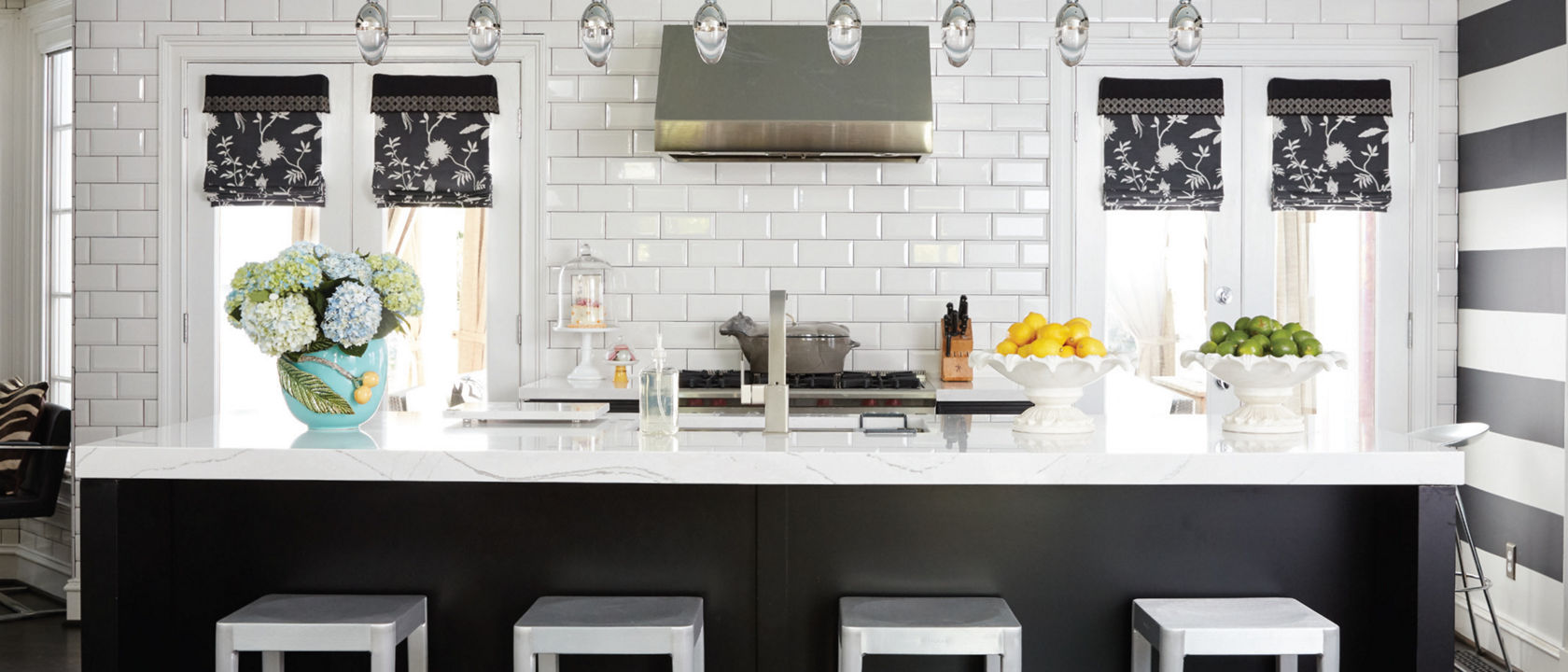 a black and white kitchen with a black center island topped with white quartz countertops and 4 white barstools, white subway tile backsplash, a black and gold range and hood and a black and white striped wall to the left. 