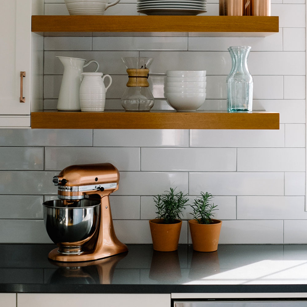 a close up of a kitchen countertop made from black quartz, with a white subway tile backsplash, open wooden shelving, white cabinets, and a rose gold stand mixer. 