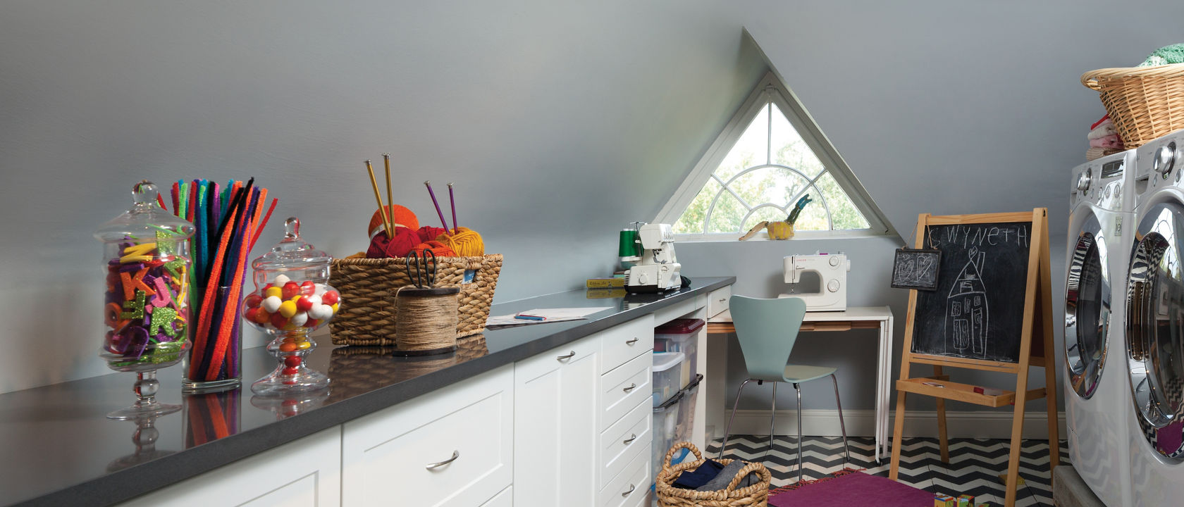 A laundry room in an attic with white cabinets, black quartz countertops, and a black and white chevron tiled floor. 