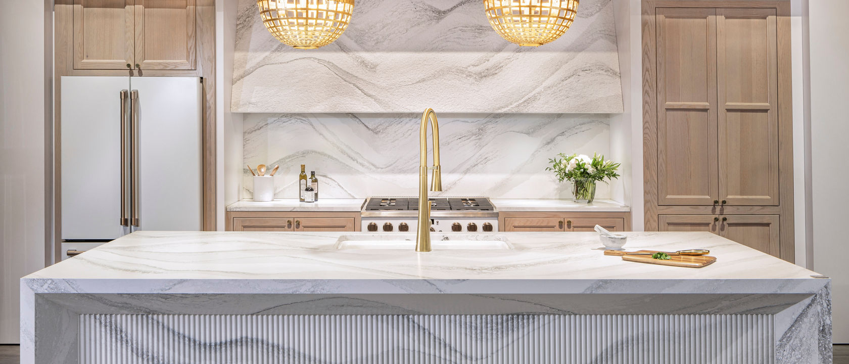 A modern kitchen with Harlow quartz double waterfall island with an island sink, matching quartz backsplash, and light wooden cabinets.