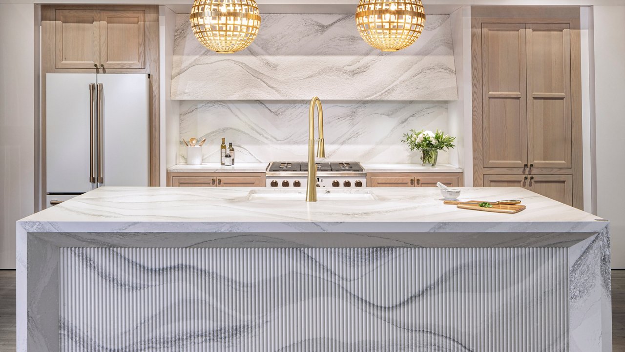 A modern kitchen with Harlow quartz double waterfall island with an island sink, matching quartz backsplash, and light wooden cabinets.