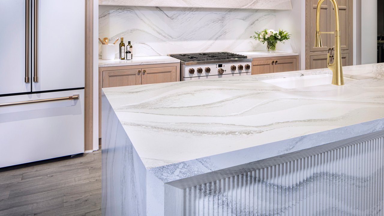 a white kitchen with unique double waterfall edge quartz island with textured front, gold accented faucet, matching quartz backsplash, and modern white appliances.