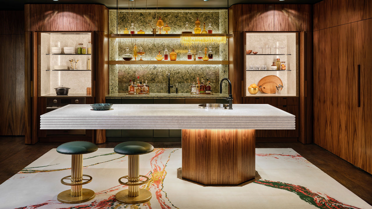 A modern bar made from white quartz with a tree trunk-inspired base, two bar stools, and open shelving with a green quartz backsplash as well as bold marbled flooring