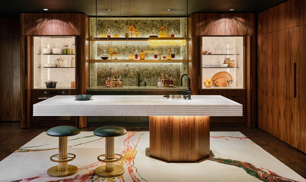 A modern bar made from white quartz with a tree trunk-inspired base, two bar stools, and open shelving with a green quartz backsplash as well as bold marbled flooring