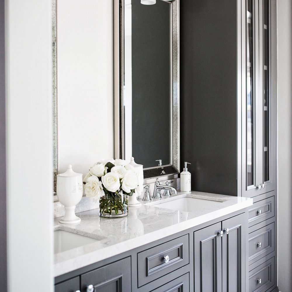A luxurious bathroom with a gray vanity, floor-to-ceiling shelving on the right side, two sinks with silver faucets, two mirrors, and lots of natural light