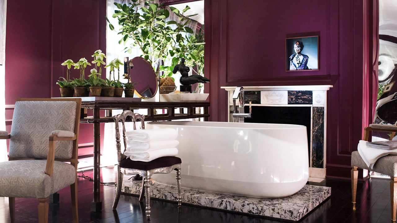 a luxurious bathroom with a white tub on tops of a quartz slab, wine red walls, elegant furniture, and lots of plants.