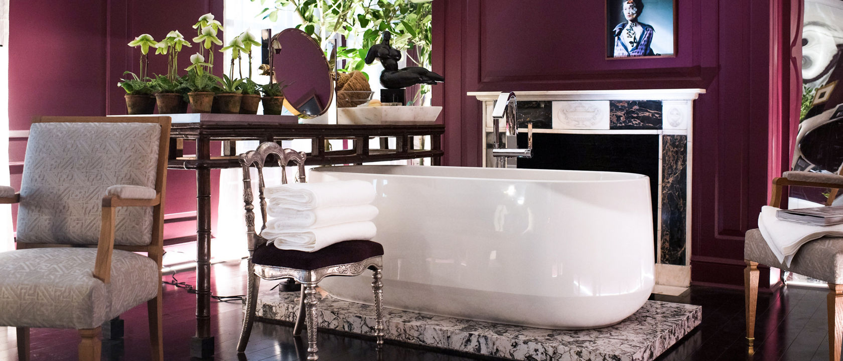 a luxurious bathroom with a white tub on tops of a quartz slab, wine red walls, elegant furniture, and lots of plants.