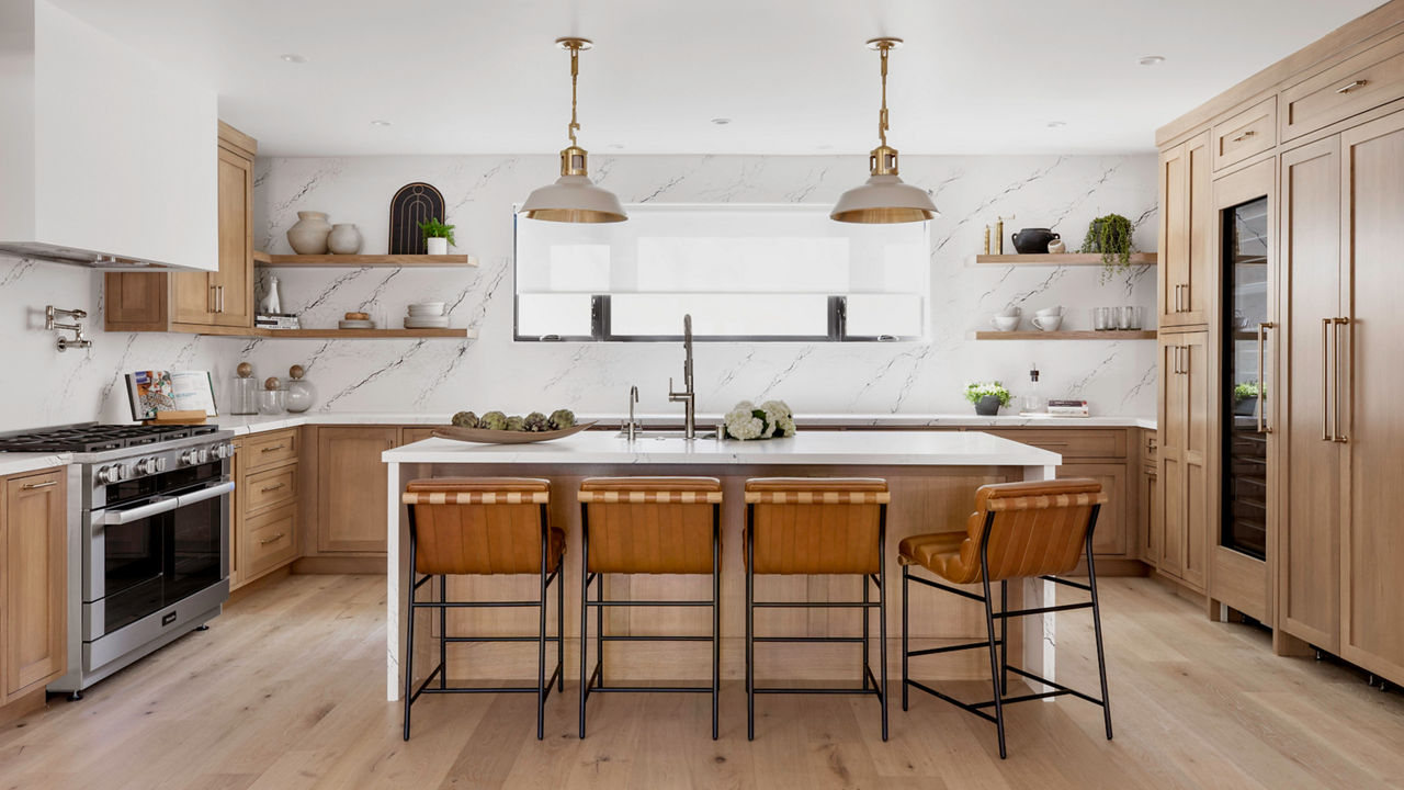 A stunning kitchen with light stained wooden cabinets, a double-waterfall edged island made from white quartz, matching quartz backsplash throughout the kitchen, and modern appliances.