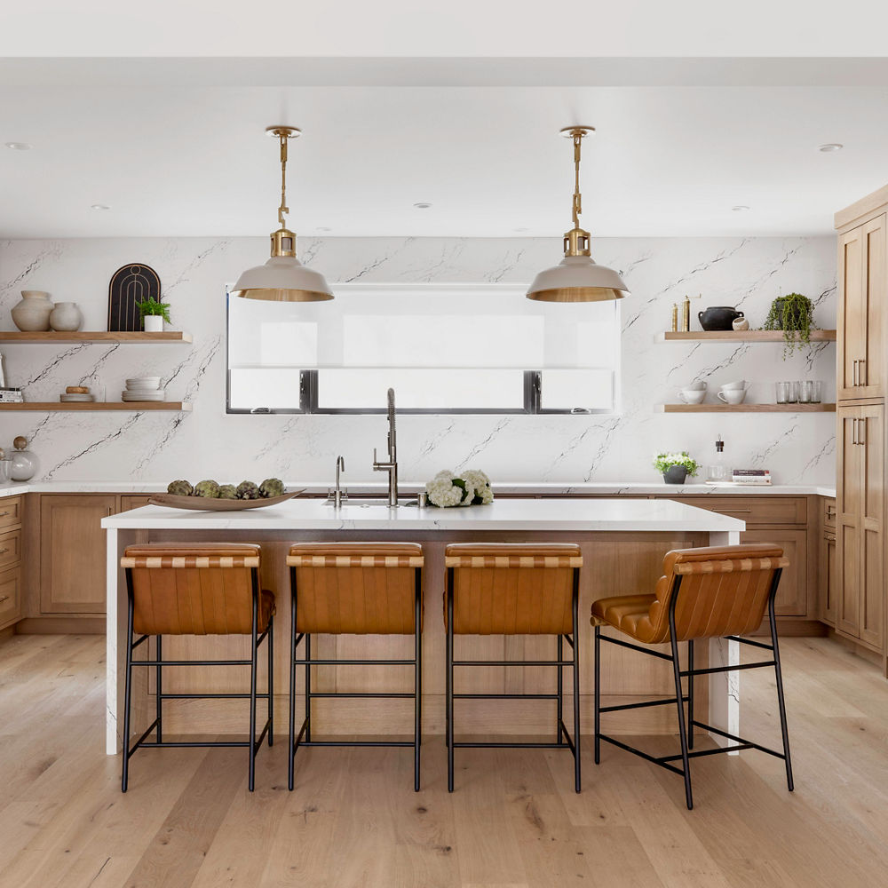 A stunning kitchen with light stained wooden cabinets, a double-waterfall edged island made from white quartz, matching quartz backsplash throughout the kitchen, and modern appliances.