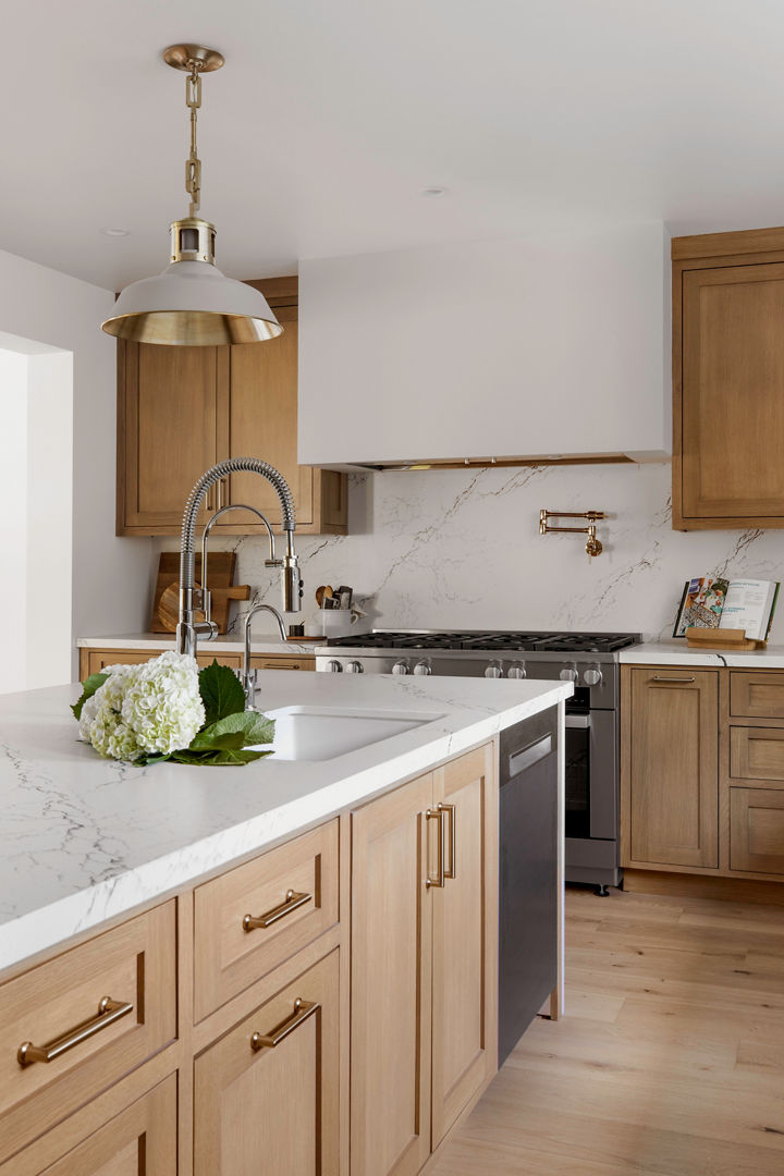 A stunning kitchen with light stained wooden cabinets, a double-waterfall edged island made from white quartz, matching quartz backsplash throughout the kitchen, and modern appliances.