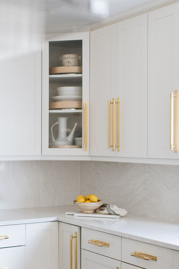 a close up of a kitchen with white cabinets with gold handles and a corner cabinet with a glass door