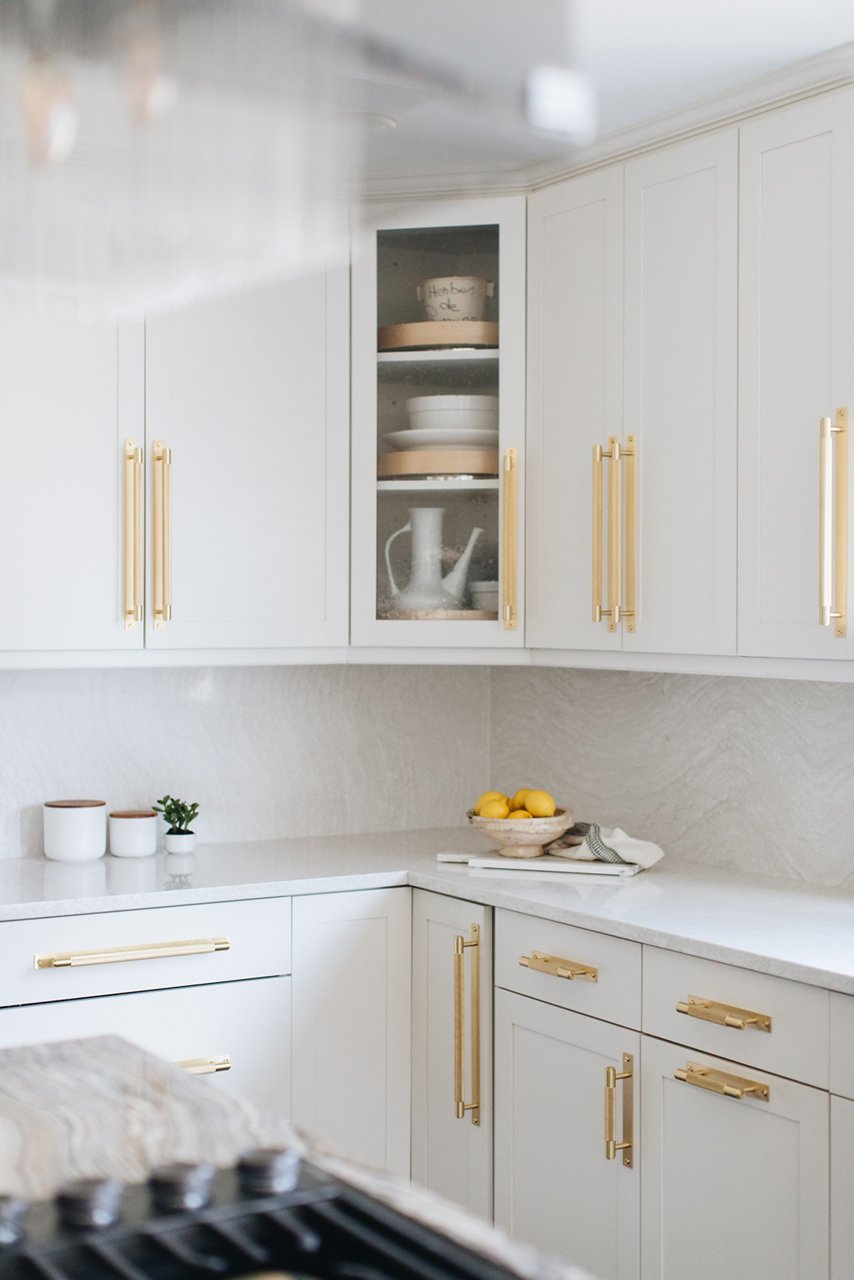 a close up of a kitchen with white cabinets with gold handles and a corner cabinet with a glass door