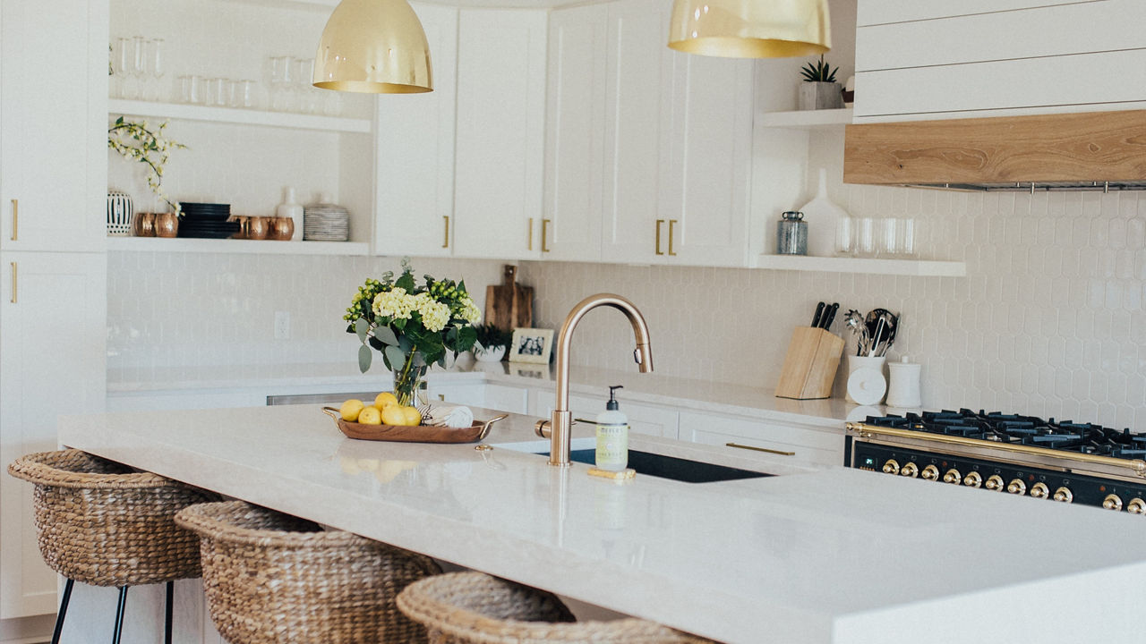 a warm kitchen with hard wood floors, white cabinets, open shelving, a black and gold range with white and wood hood, and center double waterfall-edge island with three barstools.