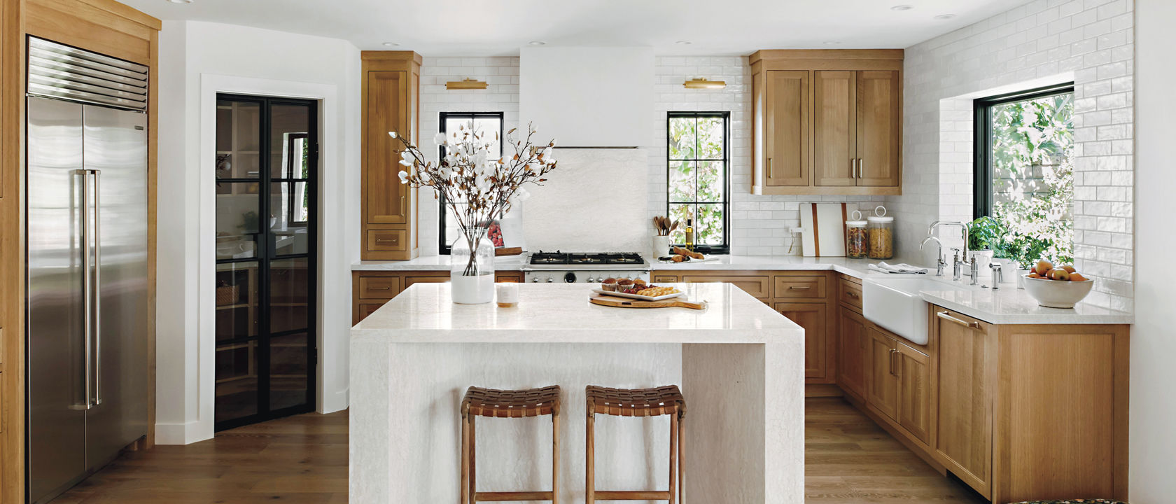a farmhouse kitchen with oak cabinets, white quartz countertops, a center double waterfall island and a gorgeous pantry with a black door made from glass.