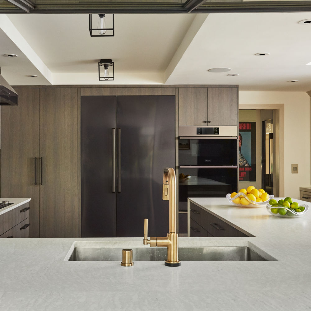 a neutral kitchen with gray-brown cabinets, matte black hardware, Cambria white quartz countertops and matching backsplash, and a sink with a golden faucet.