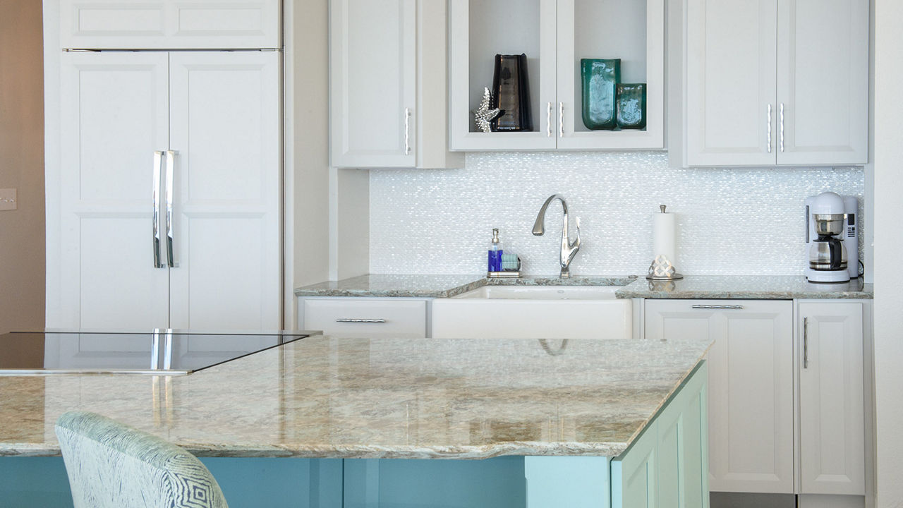 a costal inspired kitchen with white cabinets, farmhouse sink, teal center island with teal bar stools and topped with beige quartz countertops.