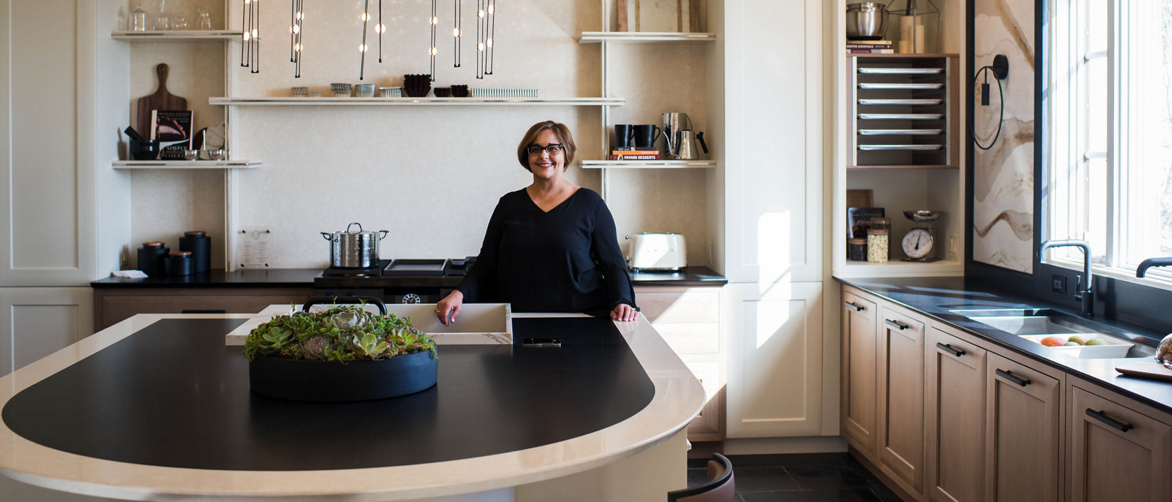 A woman stands by a round kitchen island in a kitchen with a beige and black color palette.