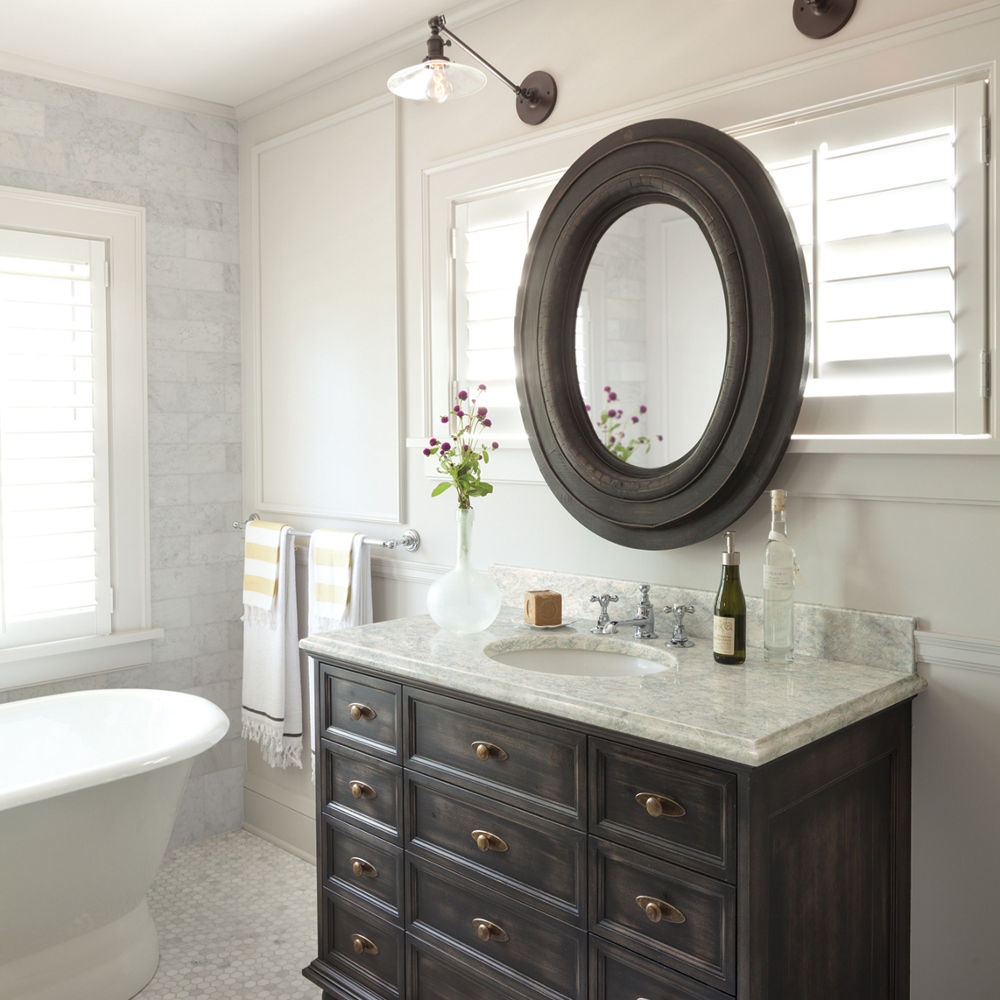 a traditional bathroom with dark wooden cabinetry, beige quartz countertops, a rounded mirror, and tiled flooring.