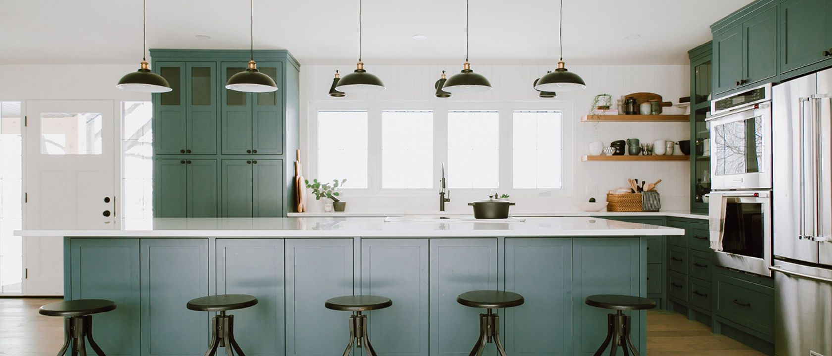 a green kitchen with green cabinets, green center island with 5 green bar stools and topped with white quartz countertops, overhead pendant lighting, 4 small windows over the sink, stainless steel appliances, and open wooden shelving