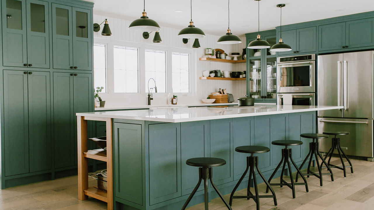 a green kitchen with green cabinets, green center island with 5 green bar stools and topped with white quartz countertops, overhead pendant lighting, 4 small windows over the sink, stainless steel appliances, and open wooden shelving