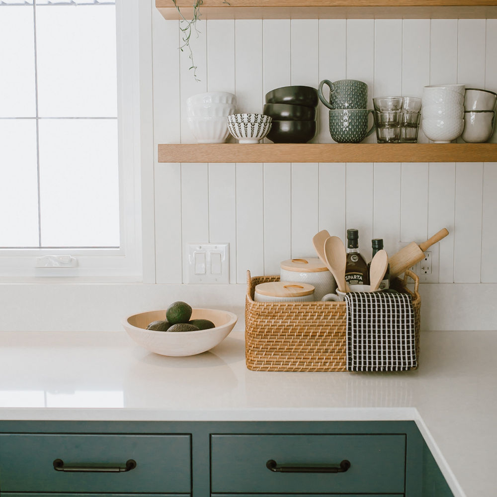 a green kitchen with green lower cabinets, white quartz countertops, open wooden shelving, and various kitchen appliances and decor on display.