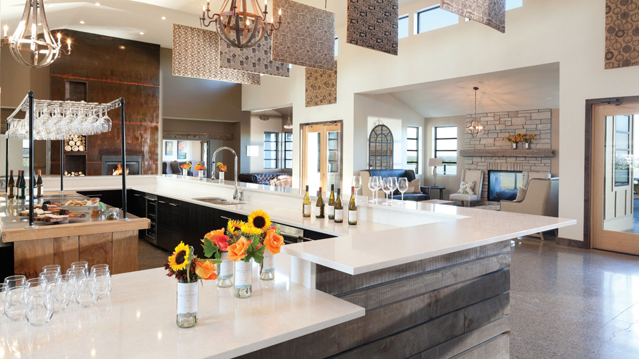 A stunning winery bar with wooden shiplap, white quartz countertops, hanging wine glasses, industrial style lighting, and a cozy seating area with a fireplace. 
